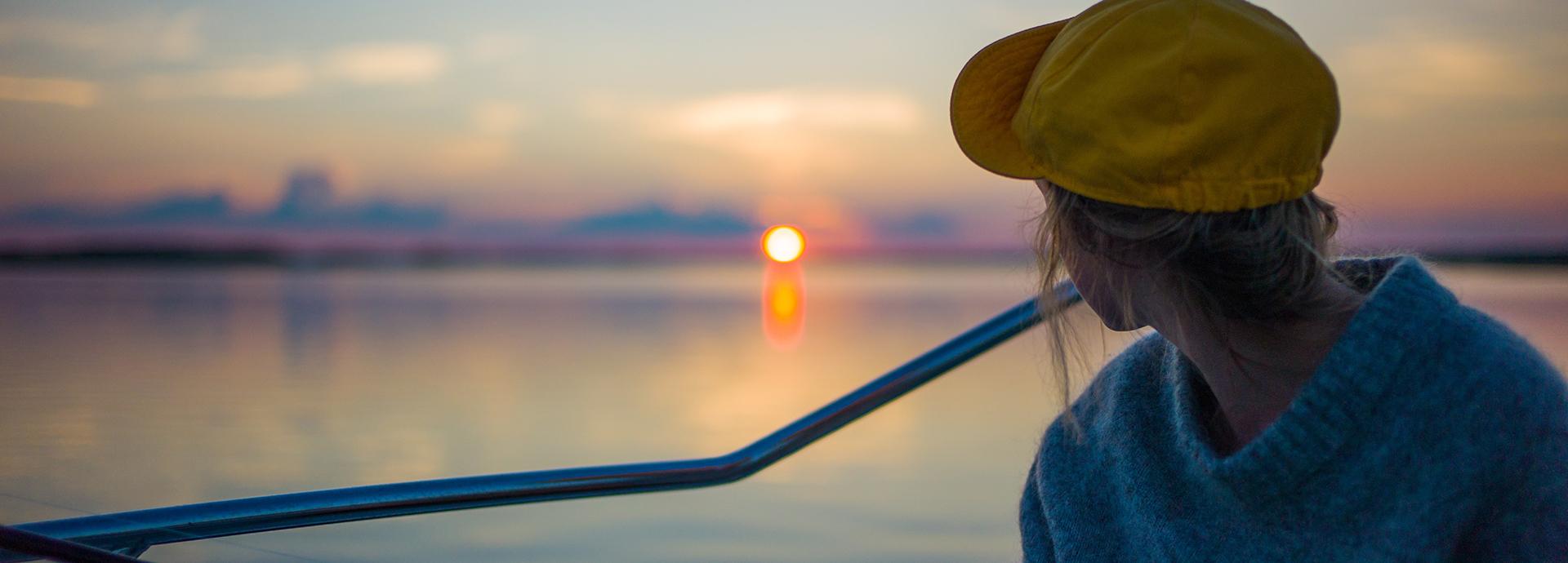 A person wearing a yellow cap sitting a boat looking at a sun setting behind a lake