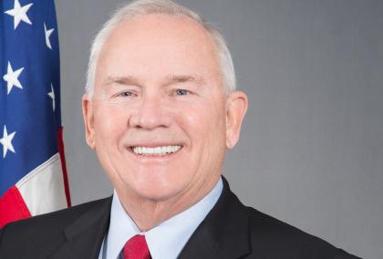 Ambassador Robert F. Pence in front of U.S. flag and grey background.