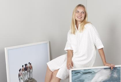 Lotta Lemetti sitting and smiling among her photos. She is wearing a white dress and is in a white room.