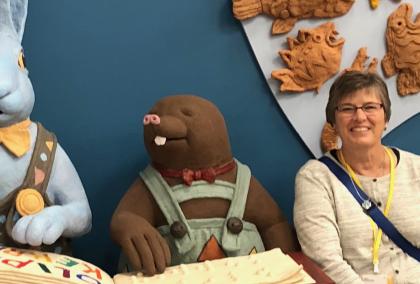 Fulbright Specialist Martha Elford sitting and smiling in front of a dark blue background. There are two large animal characters next to her: a light blue rabbit with a yellow bow tie and brown mole with a red bow tie and green overalls. The animals are reading a big story book.