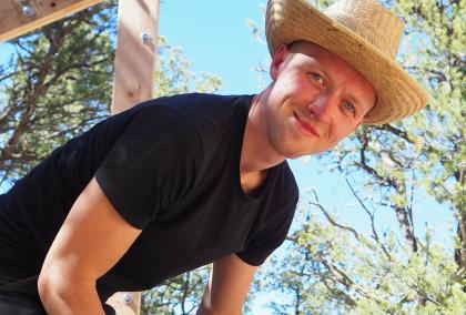 Pekko Sangi smiling at the camera, wearing a straw hat. The background shows clear blue sky and some trees.