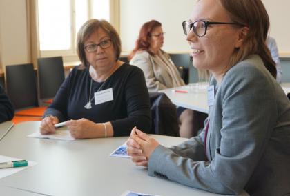 Workshop participants, two women, one of which is talking and other one is listening. They are sitting around a white round table.