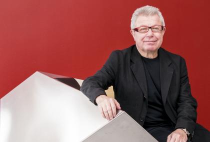 Architect Daniel Libeskind sitting on a geometrical shape with a deep red wall in the background