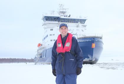 William Woityra smiling, standing on a frozen sea in front of Polaris ice breaker