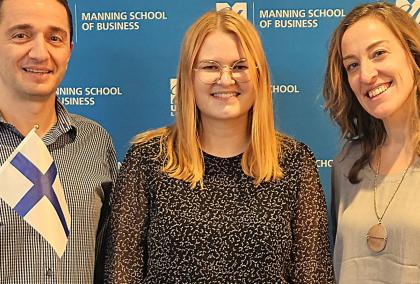 M. Berk Talays, Katariina Sorvari and Mujde Yuksel in front of Manning School of Business logo wall. Berk and Mujde are holding Finnish flags.