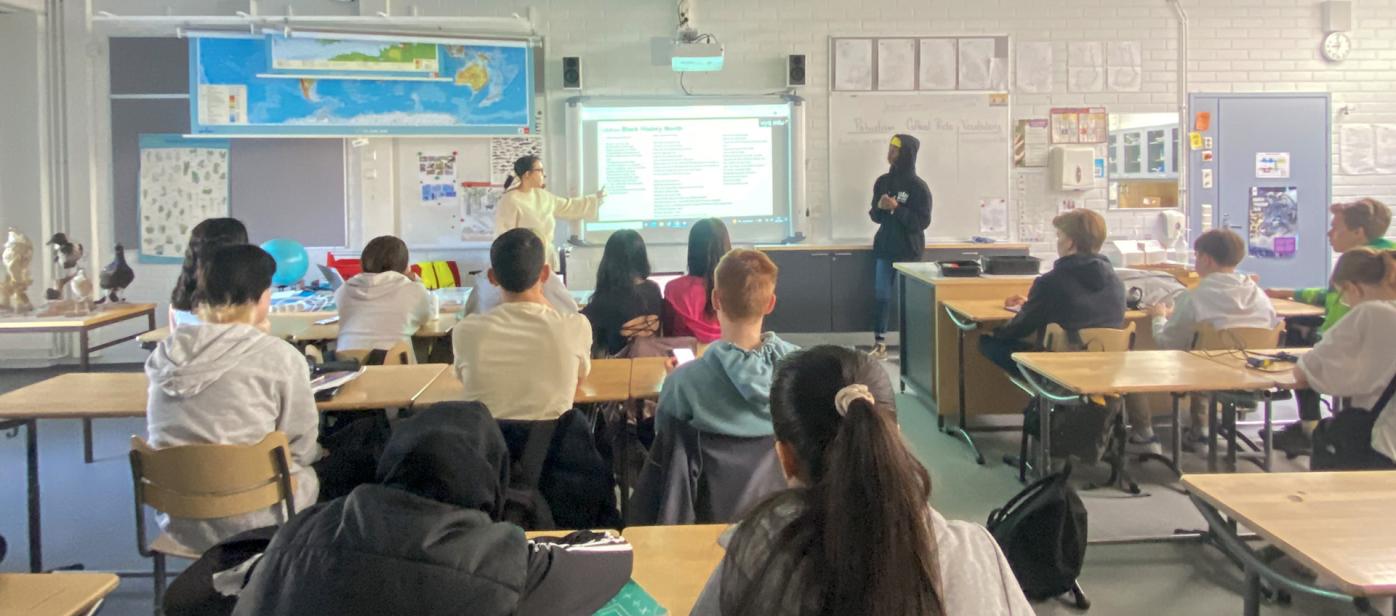 Carolyn Streets presenting to students in a classroom. Photo is taken from the back of the room showing backs of students. Carolyn is in the front, pointing at a presentation.