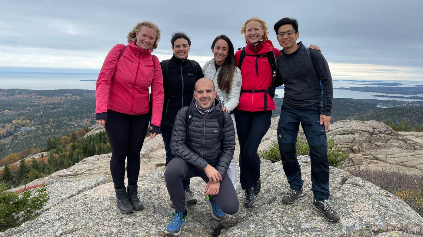 Six people in hiking gear on top of a hill smiling at the camera. Behind them opens a vast scenery with forests and lakes.