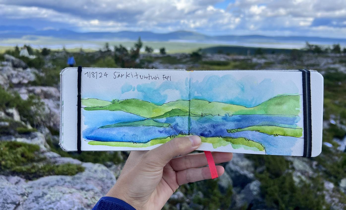 Jenny holding a small note book with a water color painting of the scenery in front of her from the Särkitunturi fell in Lapland.