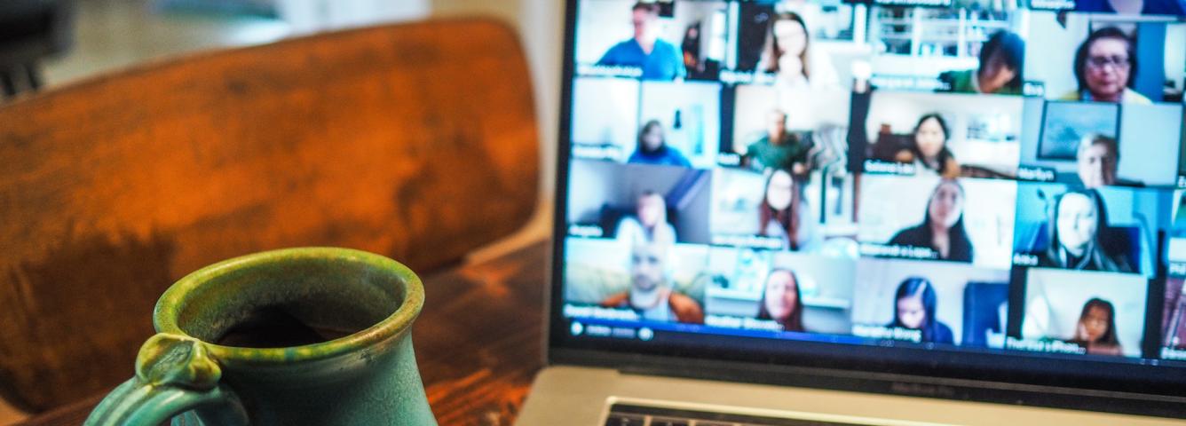 Laptop with a Zoom call gallery view with people with their cameras on and a teal colored coffee cup on the left of the photo. The cup is in focus and the laptop screen is blurry.