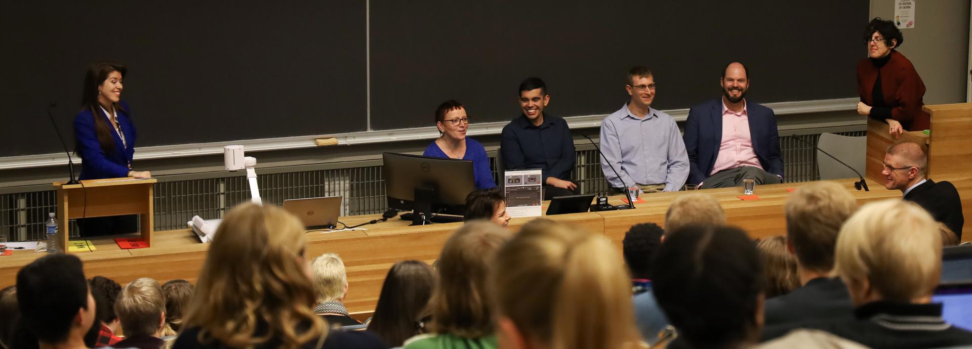 A full auditorium listening to a panel discussion. Five U.S. Fulbrighters are smiling and laughing behind a desk with a moderator smiling at them left of them.