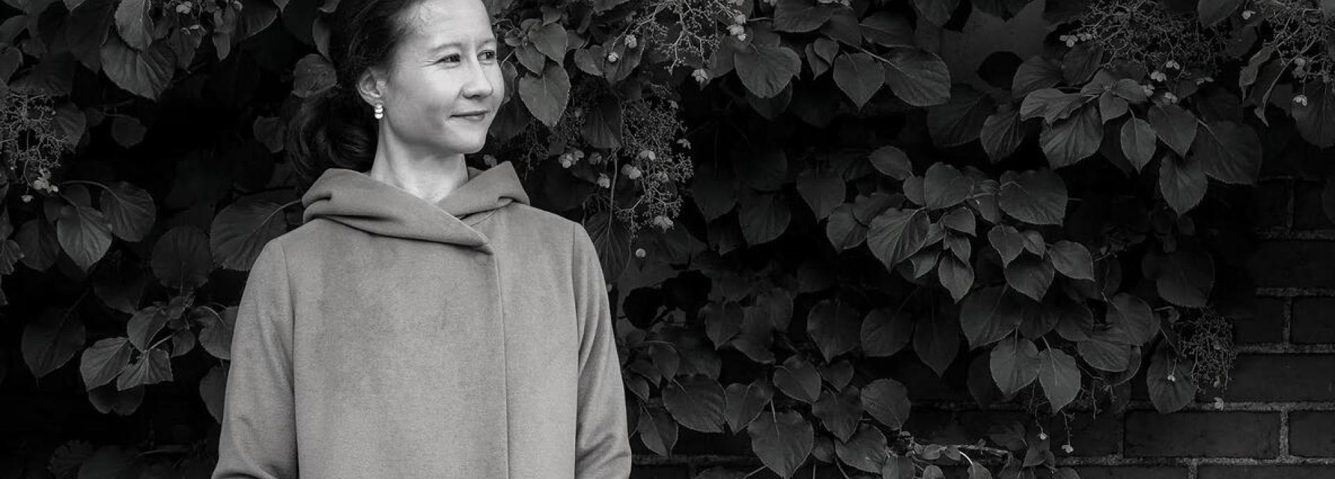 Black and white photo of Liesl Yamaguchi taken in front of a brick wall with plants hanging on it. Liesl is wearing a hoodie and she looks to her left.