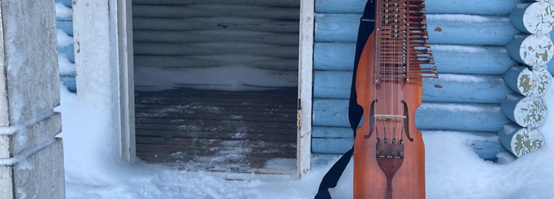 Nyckelharpa leaning against light blue wall of a Finnish wooden house. It's winter, and there is snow on the porch. The door next to the nyckelharpa is open