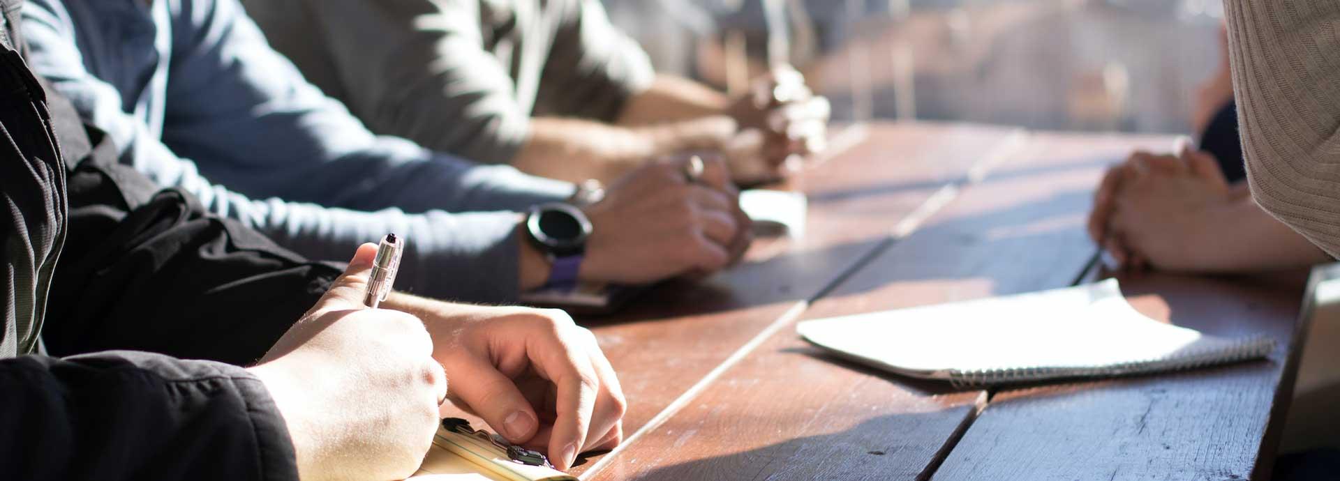 Arms and hands of four people who are sitting around a table having a meeting. One of the people is holding a pen and writing somethign on a paper.