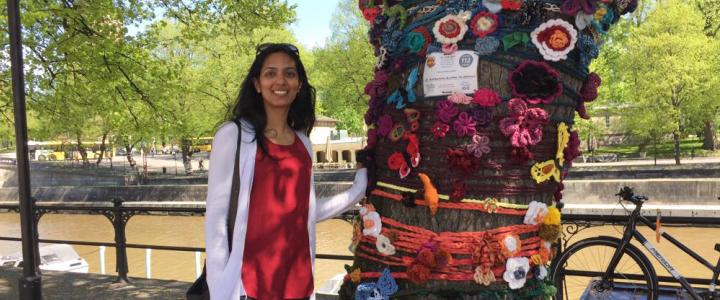 Avanti Chajed standing next to a tree, which is covered in knitted patterns. The photo is taken on a sunny day next to River Aura in Turku, Finland.