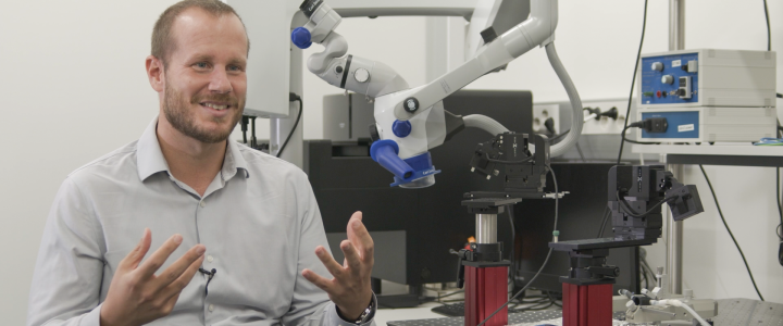 Nelson Totah sitting in front of a microscope in a laboraty. He is looking at the interviewer, explaining something with also using his hands.