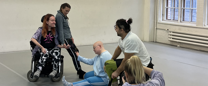 A photographer captures one dancer leaning against another in a wheelchair and one more kneeling on the ground while another wheelchair dancer moves behind.