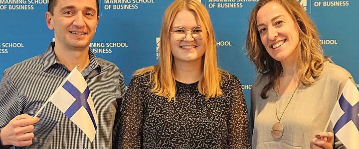 M. Berk Talays, Katariina Sorvari and Mujde Yuksel in front of Manning School of Business logo wall. Berk and Mujde are holding Finnish flags.