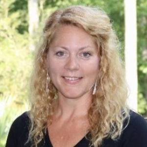 Person with curly shoulder-length blonde hair in a black shirt smiling in front of a nature background with trees