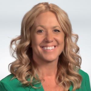 Person with shoulder-length light hair in a green jacket smiling in front of a white background