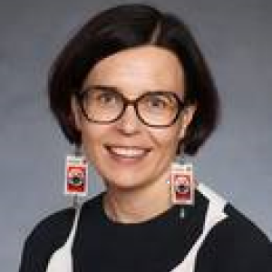 Person with black short hair, large rectangle shaped earrings and glasses smiling at the camera