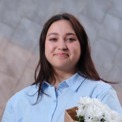 Person with shoulder-length brown hair wearing a light blue button-up shirt in front of a grey background